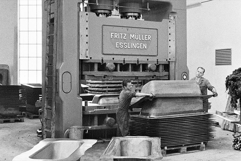 Historische Schwarz-Weiss-Aufnahme von Mitarbeitenden an einer grossen Pressemaschine beim Formen von Badewannen.