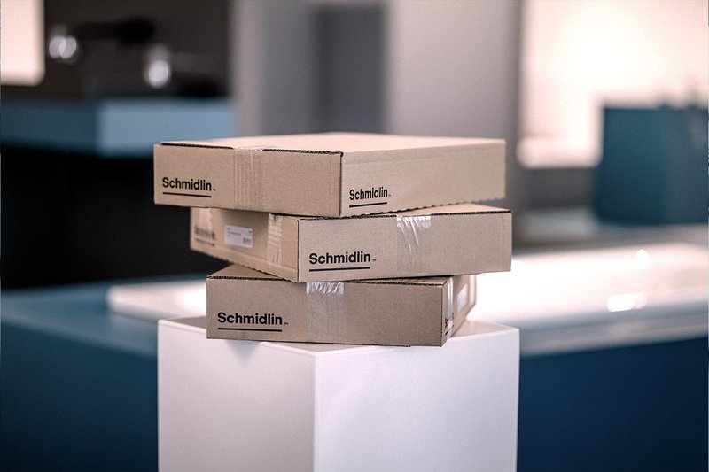 Drei gestapelte Schmidlin-Kartons auf einem weissen Podest, in modernem Innenraum mit kühlem Farblook.