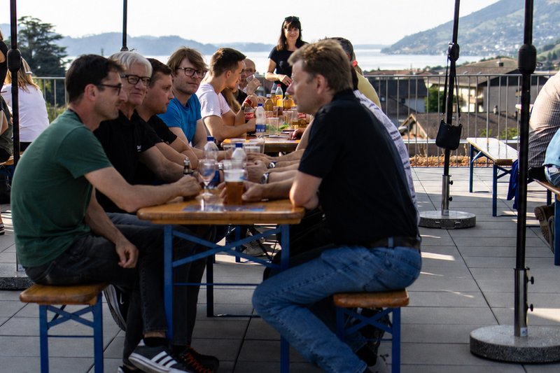 Mitarbeitende sitzen bei einem gemeinsamen Essen an langen Holztischen auf einer Terrasse mit Blick auf See und Berge.