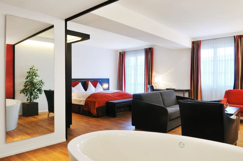 Modernes Hotelzimmer mit freistehender Badewanne, Wohnbereich und Schlafzimmer mit roten Akzenten