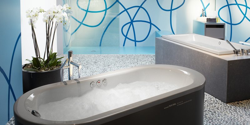 Freistehende, ovale Badewanne mit Schaumfüllung in modernem Badezimmer, daneben Orchidee und Armatur; im Hintergrund zweite Wanne vor blau gemusterter Glaswand.