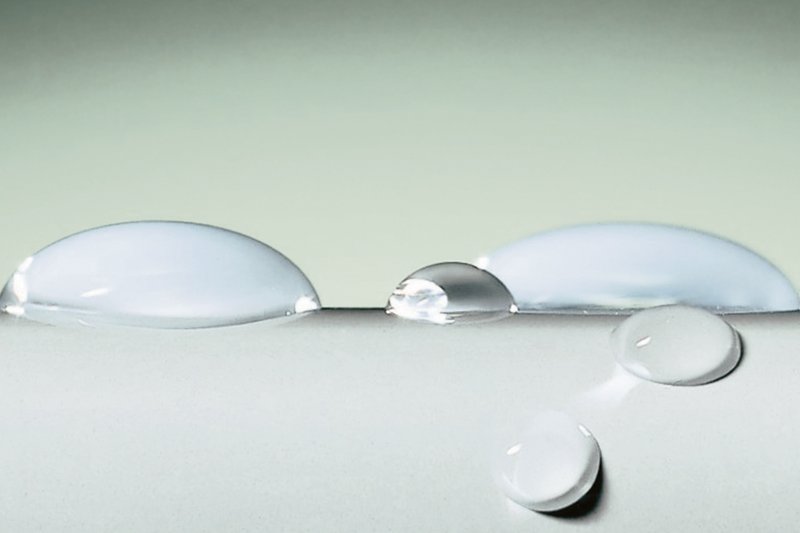 Makroaufnahme mehrerer klarer Wassertropfen auf einer glatten, hellen Oberfläche.