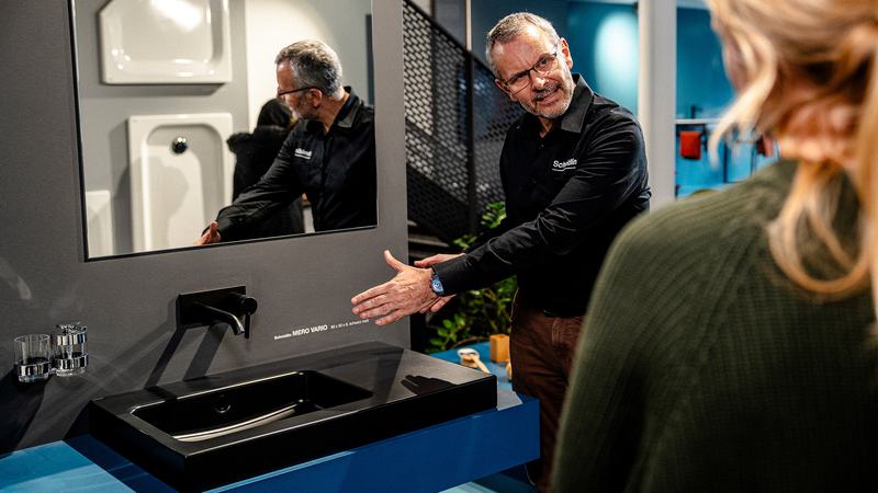 Ein Schmidlin-Mitarbeiter erklärt einer Besucherin ein schwarzes Waschbeckenmodell Schmidlin MERO VARIO in einer modernen Ausstellung mit Badprodukten.
