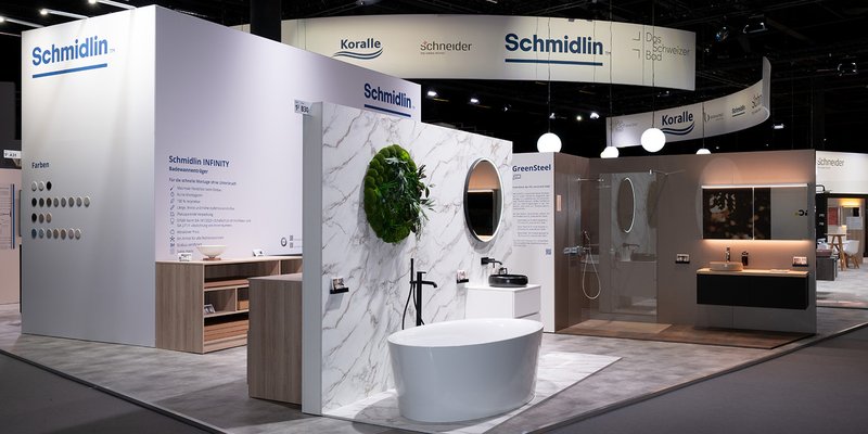 Messestand von Schmidlin auf einer Fachmesse mit freistehender Badewanne, Duschbereich, Badmöbeln und Markenlogos von Schmidlin, Koralle und Schneider.
