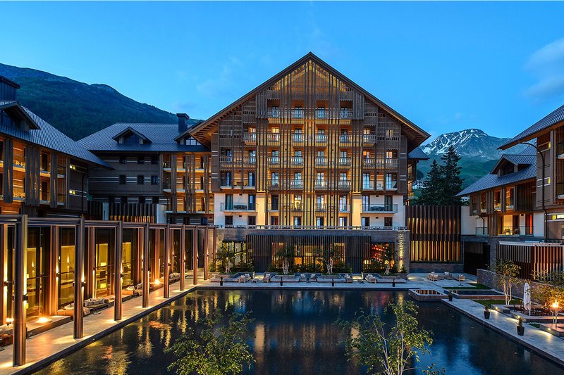 Das Chedi Andermatt bei Abenddämmerung mit beleuchtetem Innenhof, Wasserbecken und alpiner Bergkulisse.